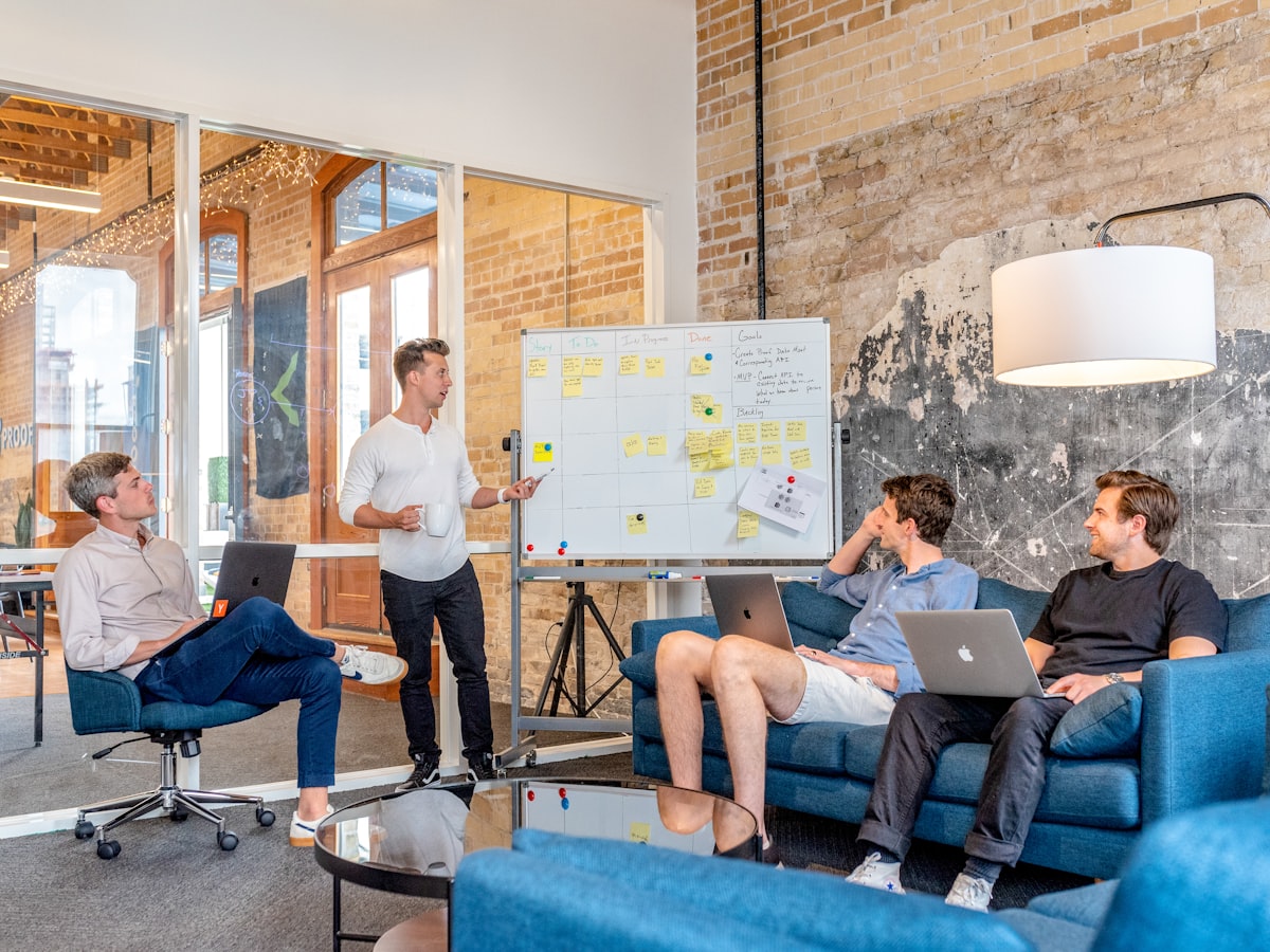 Three colleagues using laptops while a teammate presents beside a whiteboard